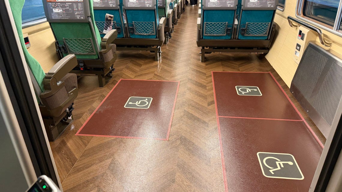 A train car interior showing two dedicated wheelchair spaces marked with large white symbols on the wood-grain floor, located adjacent to green passenger seats. A train car interior showing two dedicated wheelchair spaces marked with large white symbols on the wood-grain floor, located adjacent to green passenger seats.