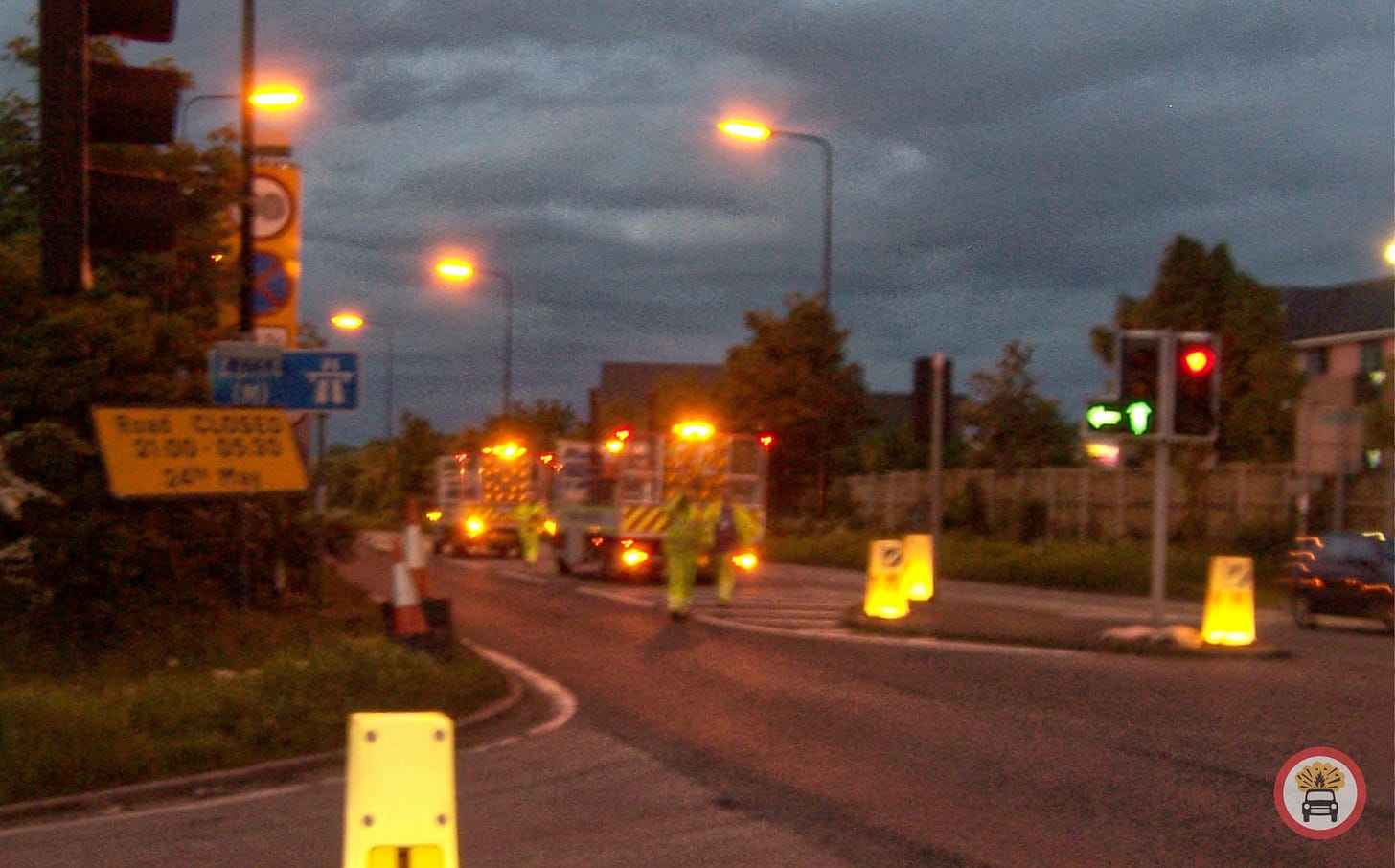 Slightly blurry photograph, taken at twilight, of the entrance to the A6144(M) with the blue motorway sign visible on the left. A works van with flashing amber beacons is stopped on the road and several workmen in hi-viz clothing can be seen moving across the road, evidently about to close it using traffic cones that are stacked up at the kerbside Slightly blurry photograph, taken at twilight, of the entrance to the A6144(M) with the blue motorway sign visible on the left. A works van with flashing amber beacons is stopped on the road and several workmen in hi-viz clothing can be seen moving across the road, evidently about to close it using traffic cones that are stacked up at the kerbside