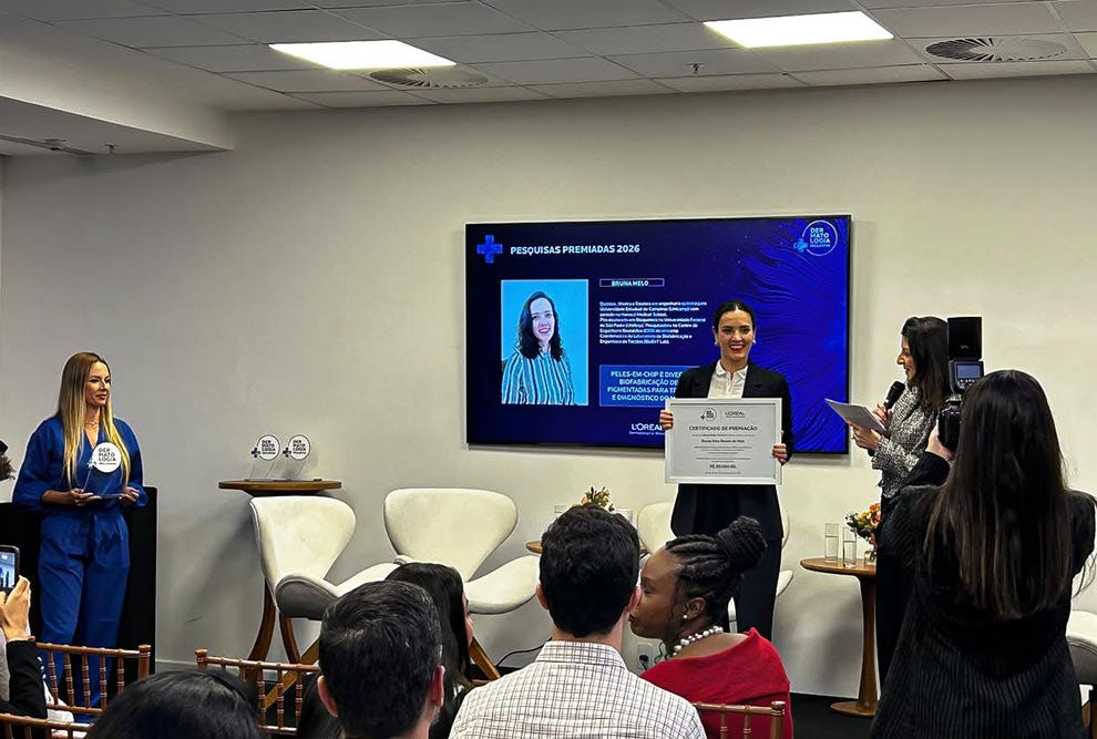 Evento em ambiente corporativo com tela de apresentação azul exibindo conteúdo sobre pesquisas premiadas 2025, onde pessoa em blazer azul está à esquerda, pessoa em blazer bege segura certificado no centro, pessoa em blazer estampado com câmera está à direita, e público sentado de costas observa a cena em cadeiras dispostas no espaço, com móveis brancos e cinzas ao fundo, iluminação de teto fluorescente e decoração minimalista.