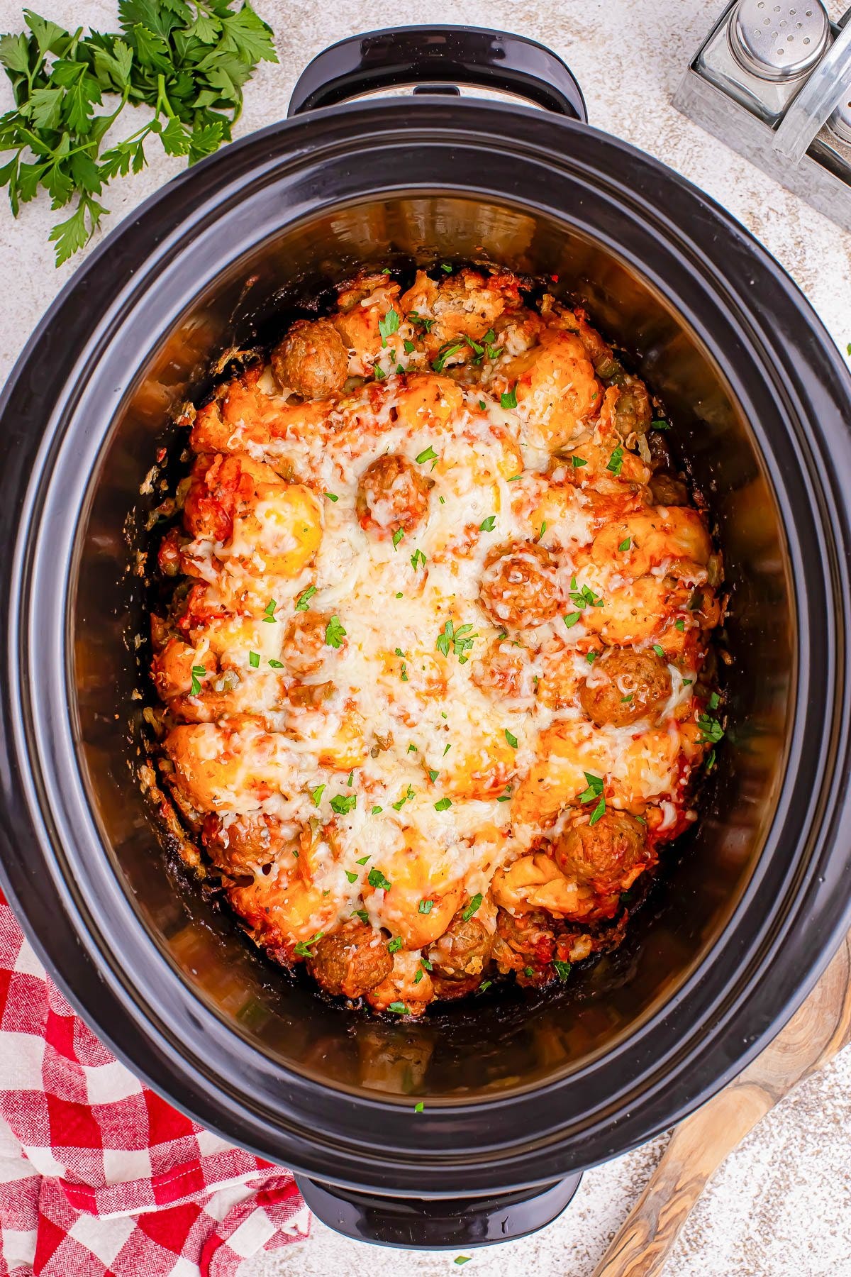A slow cooker containing meatballs in a tomato sauce, topped with melted cheese and garnished with chopped herbs.