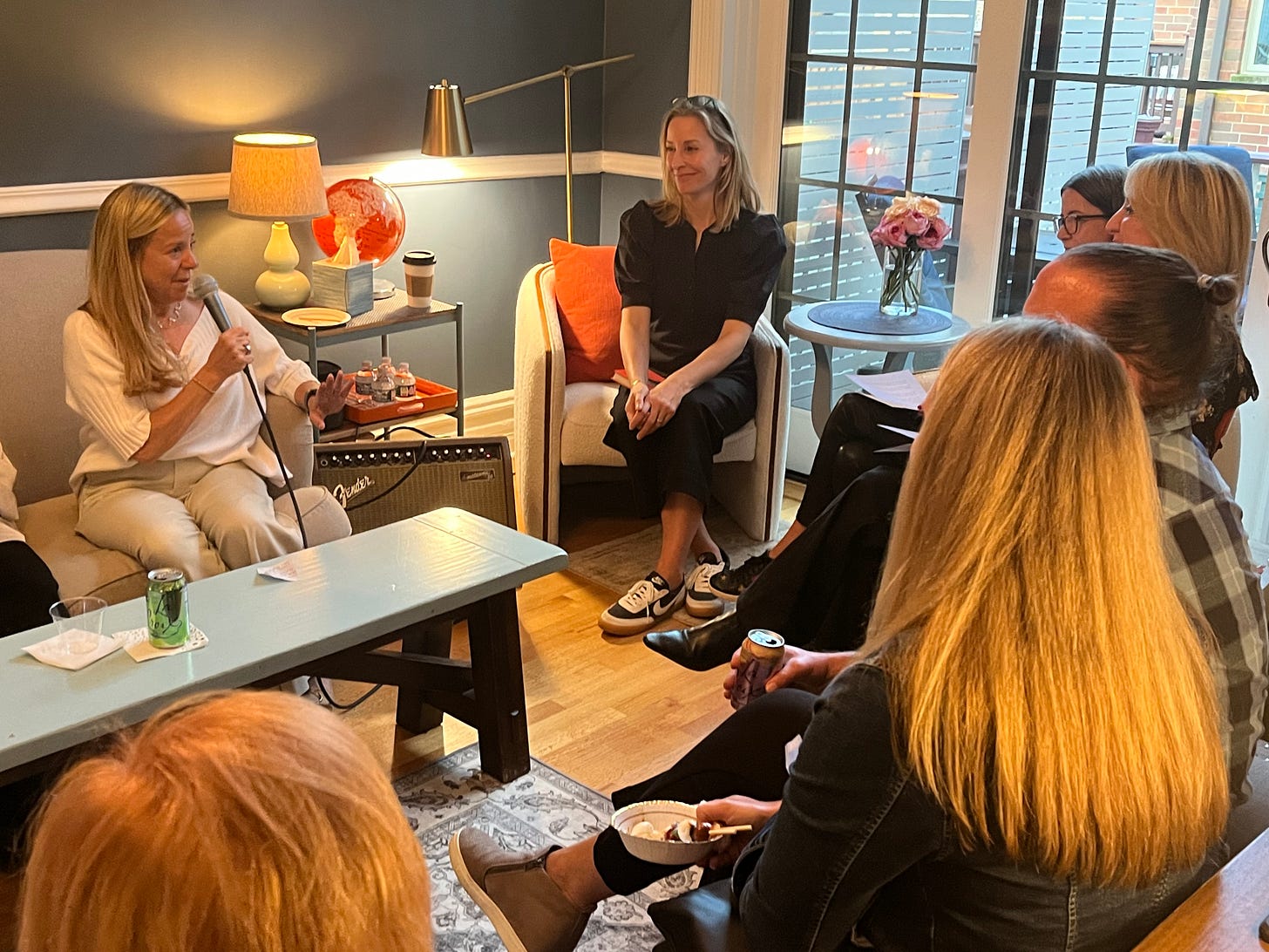 Blonde woman with microphone sits on couch speaking to a group of other women. Walls are blue. Pops of orange everywhere. 
