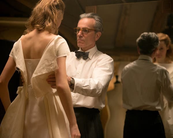 A man in a white shirt and bow tie adjusts the formal beige dress of a woman with blonde hair.