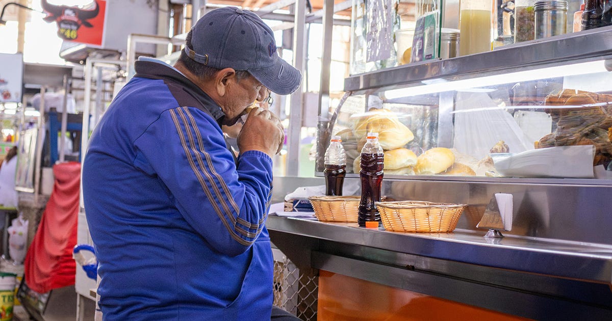 Fotografia colorida, em plano médio e de perfil, de um homem com sobrepeso, usando uma jaqueta azul, comendo em um balcão de lanchonete. Fotografia colorida, em plano médio e de perfil, de um homem com sobrepeso, usando uma jaqueta azul, comendo em um balcão de lanchonete.