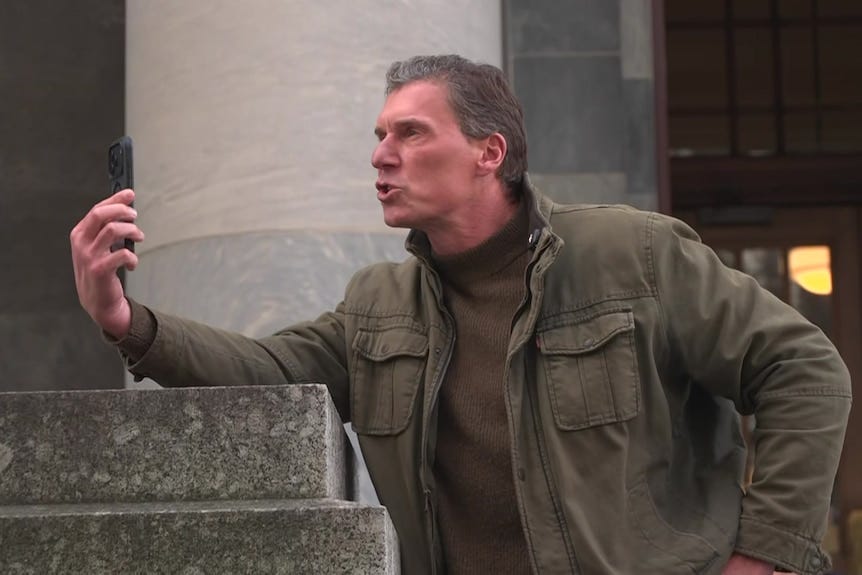 A man wearing an army-green jacket and brown turtle neck records a video in front of a stone building