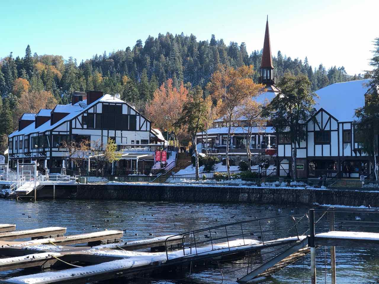 Lake Arrowhead Village along the waterfront, featuring Tudor-style buildings with white and dark timber exteriors and a tall church-like steeple. Snow lightly covers rooftops and docks along the lake, while forested hills rise in the background under a clear blue sky.