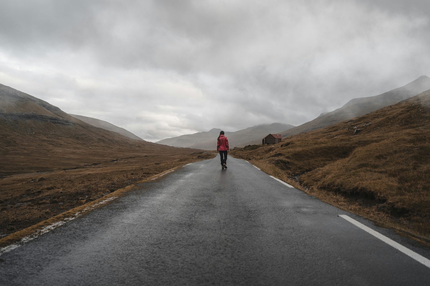 A solitary person in a red jacket walks down a wet, winding road through a misty, mountainous landscape. The sky is overcast, and the surrounding hills are covered in muted brown grass. A solitary person in a red jacket walks down a wet, winding road through a misty, mountainous landscape. The sky is overcast, and the surrounding hills are covered in muted brown grass.