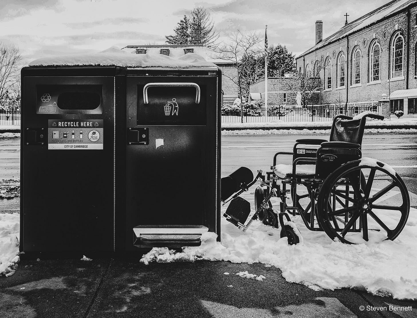 Black-and-white photograph: a manual wheelchair, snow settled on its seat and leg rests still attached, parked in neat alignment beside two large City of Cambridge trash and recycling bins on a snowy sidewalk. Across the street, a brick church and an American flag. Black-and-white photograph: a manual wheelchair, snow settled on its seat and leg rests still attached, parked in neat alignment beside two large City of Cambridge trash and recycling bins on a snowy sidewalk. Across the street, a brick church and an American flag.