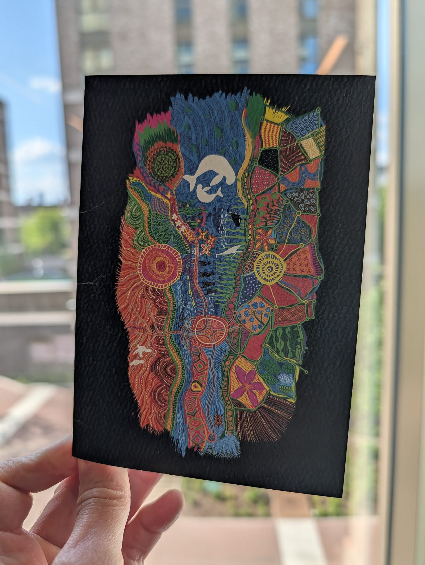 Front of a cream card featuring a striking geometric portrait in red, dark blue, green, and yellow, split down the middle showing two face halves composed of intricate patterned shapes and cultural motifs.