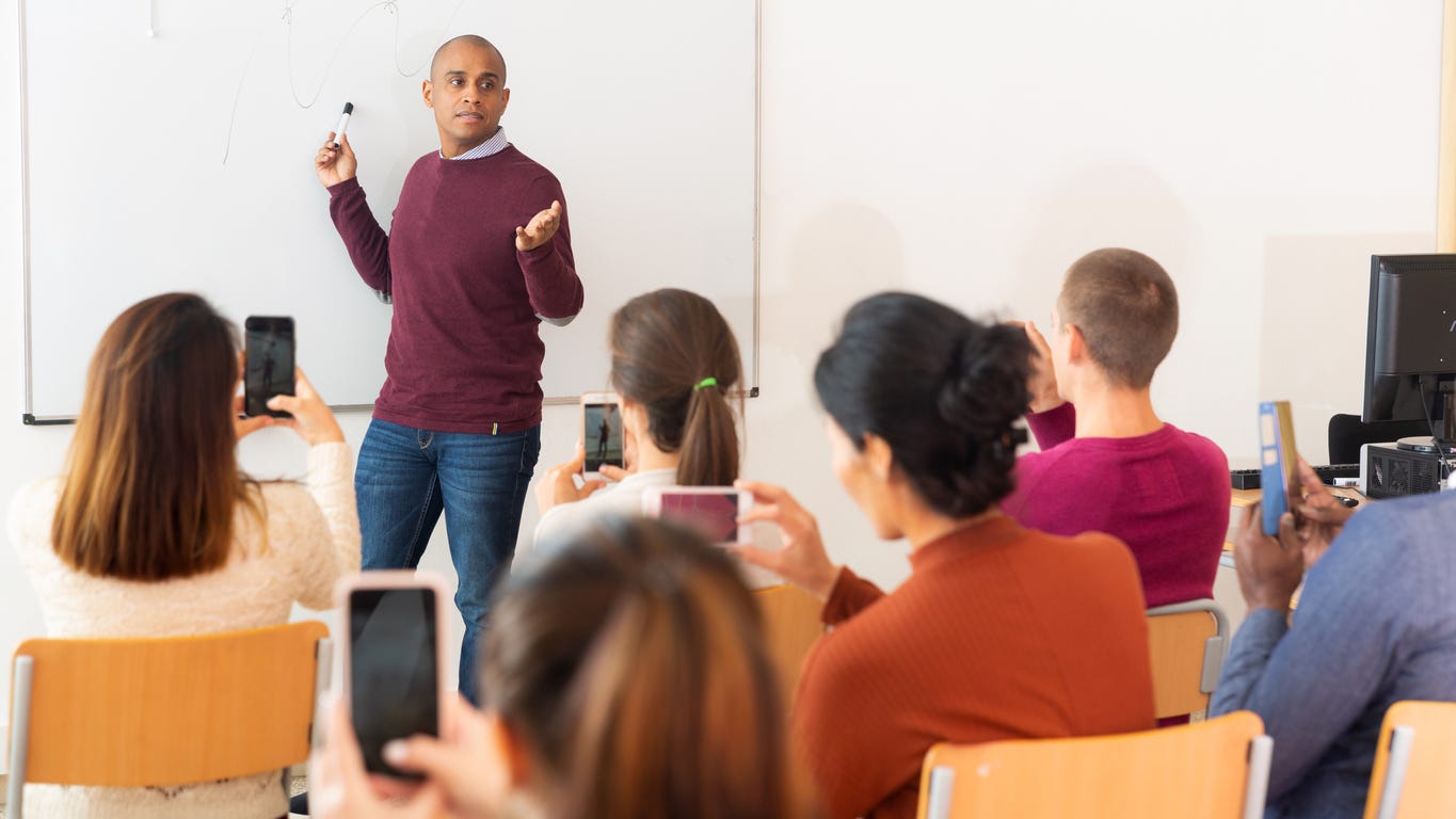 Visual showing a classroom, teaching at the whiteboard teaching, students sitting on chairs, taking pictures of the lecture using their smartphones. 