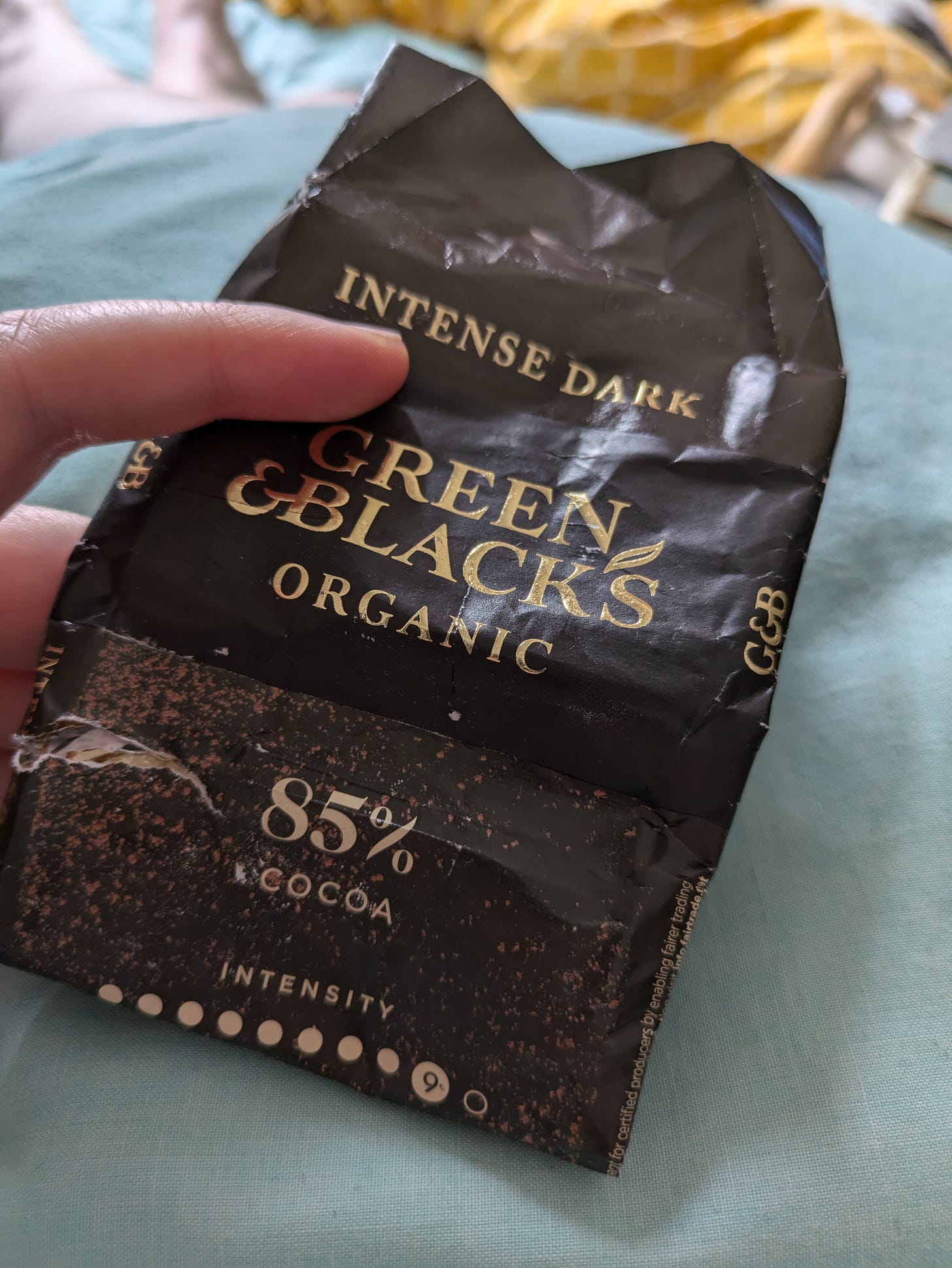 ID. A photo of me holding out a nearly finished block of Green & Blacks organic 85% dark chocolate on top of alight blue bed sheet. ID. A photo of me holding out a nearly finished block of Green & Blacks organic 85% dark chocolate on top of alight blue bed sheet.