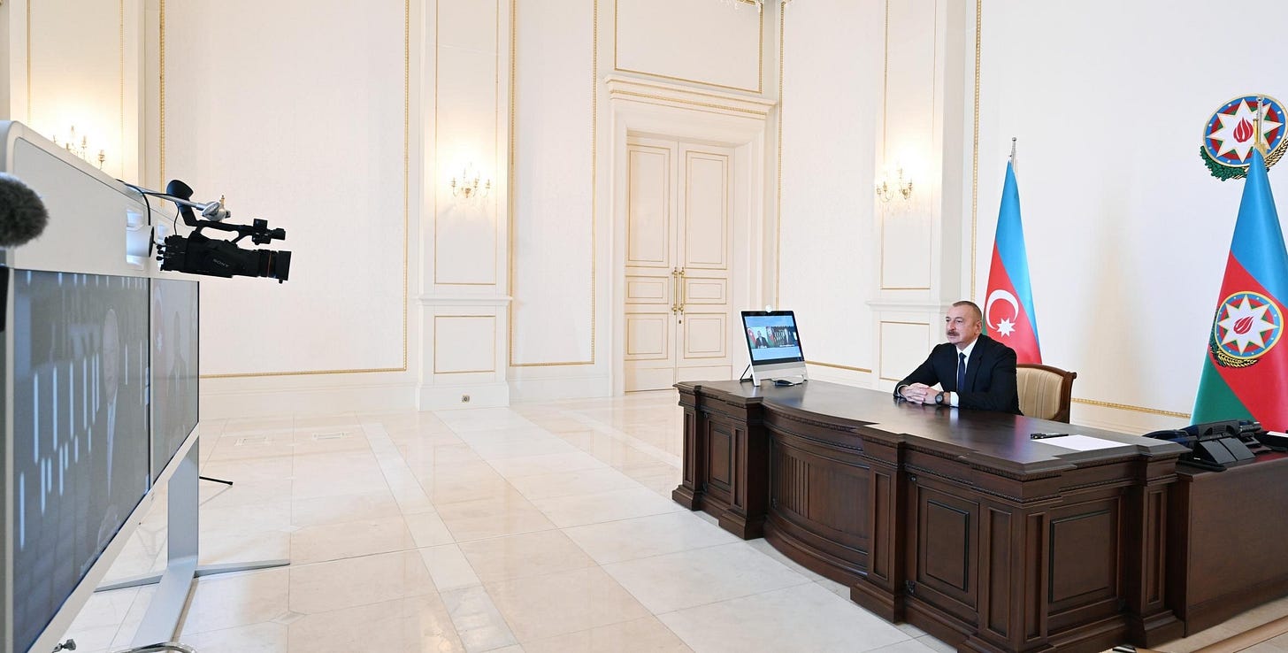 A man sits alone at a large wooden desk in a bright hall with an Azerbaijani flag and state emblem behind him, facing a television screen and a video camera on a tripod. A man sits alone at a large wooden desk in a bright hall with an Azerbaijani flag and state emblem behind him, facing a television screen and a video camera on a tripod.
