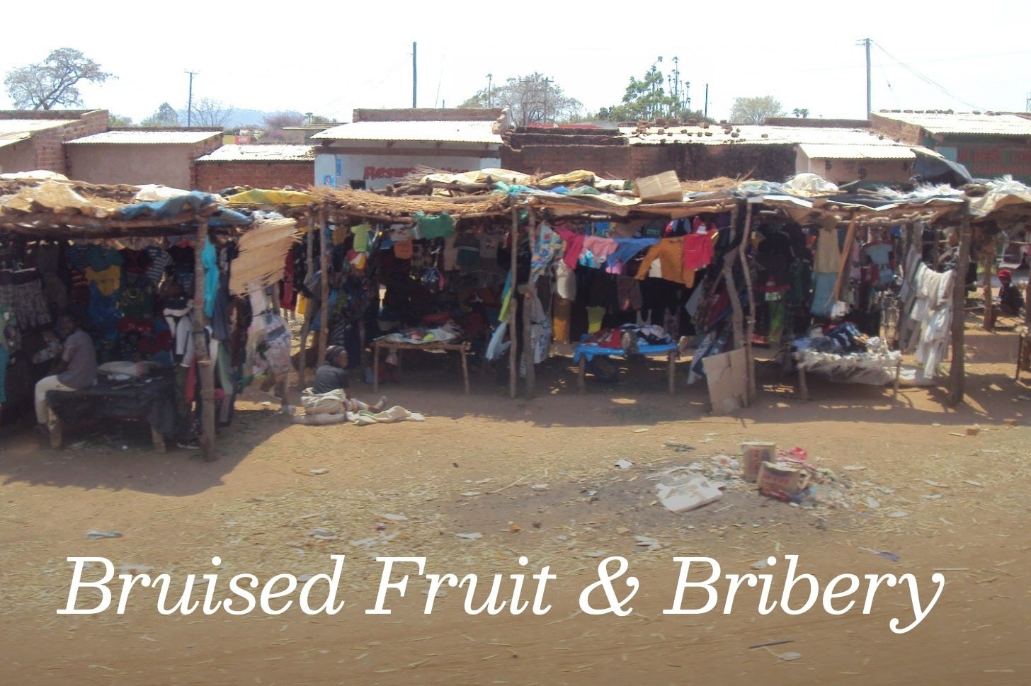 Road-side stands in rural Malawi, featuring dusty yellow road, stick buildings selling colorful clothing. Wooden huts with tin roofs are in the background.