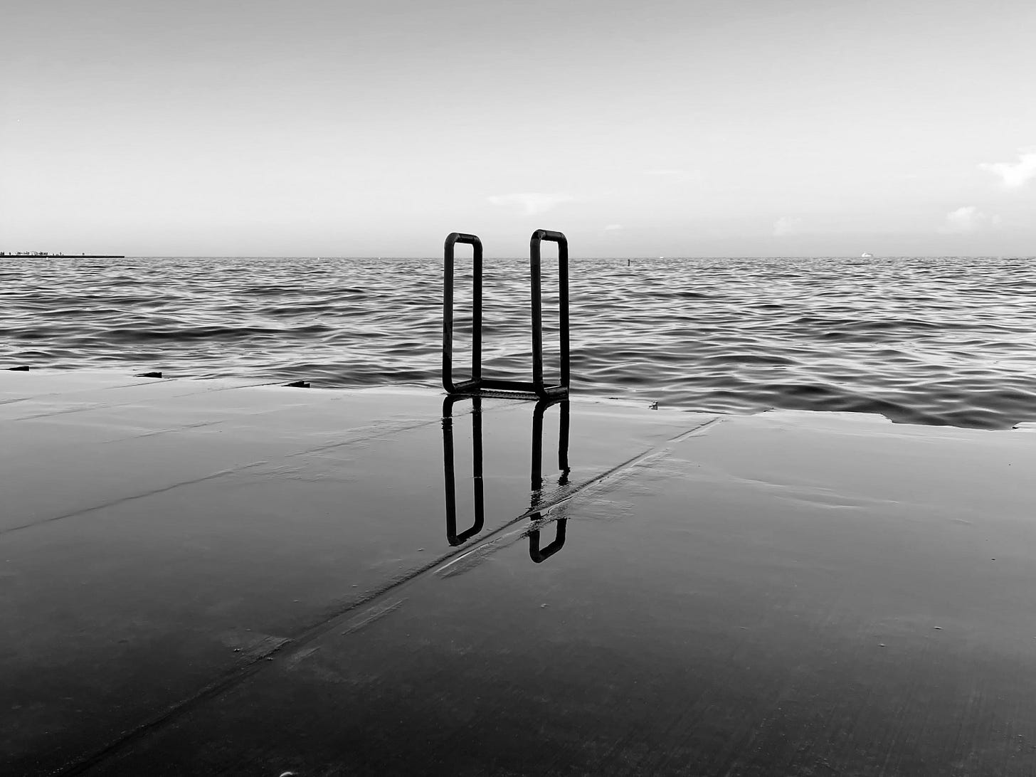 A black-and-white photo of the top of a ladder providing access to the water at a concrete-covered shore. The ladder and sky are smoothly reflected in a thin layer of water covering the concrete.