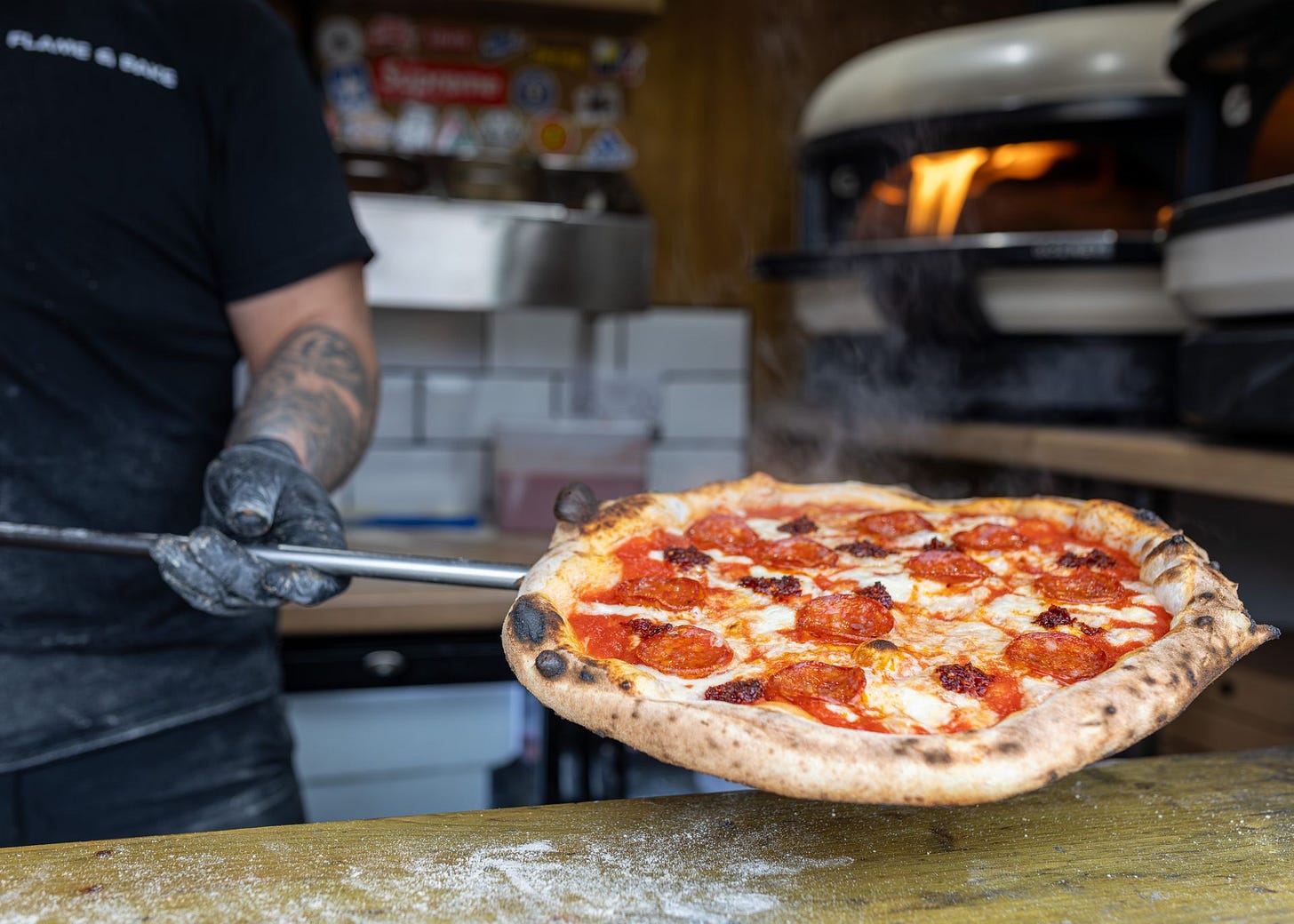 A close-up photograph of a chef holding up a freshly baked, steaming wood-fired pizza topped with melted mozzarella, pepperoni, and spicy nduja sausage on a metal pizza peel. In the background, flames roar inside a modern commercial pizza oven.