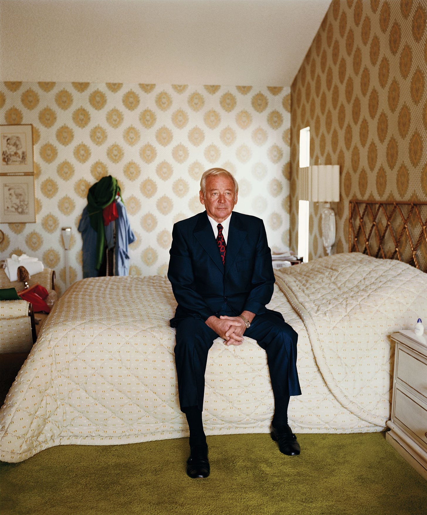 Portrait of man in suit sitting on bed in room with wallpaper and green carpet.