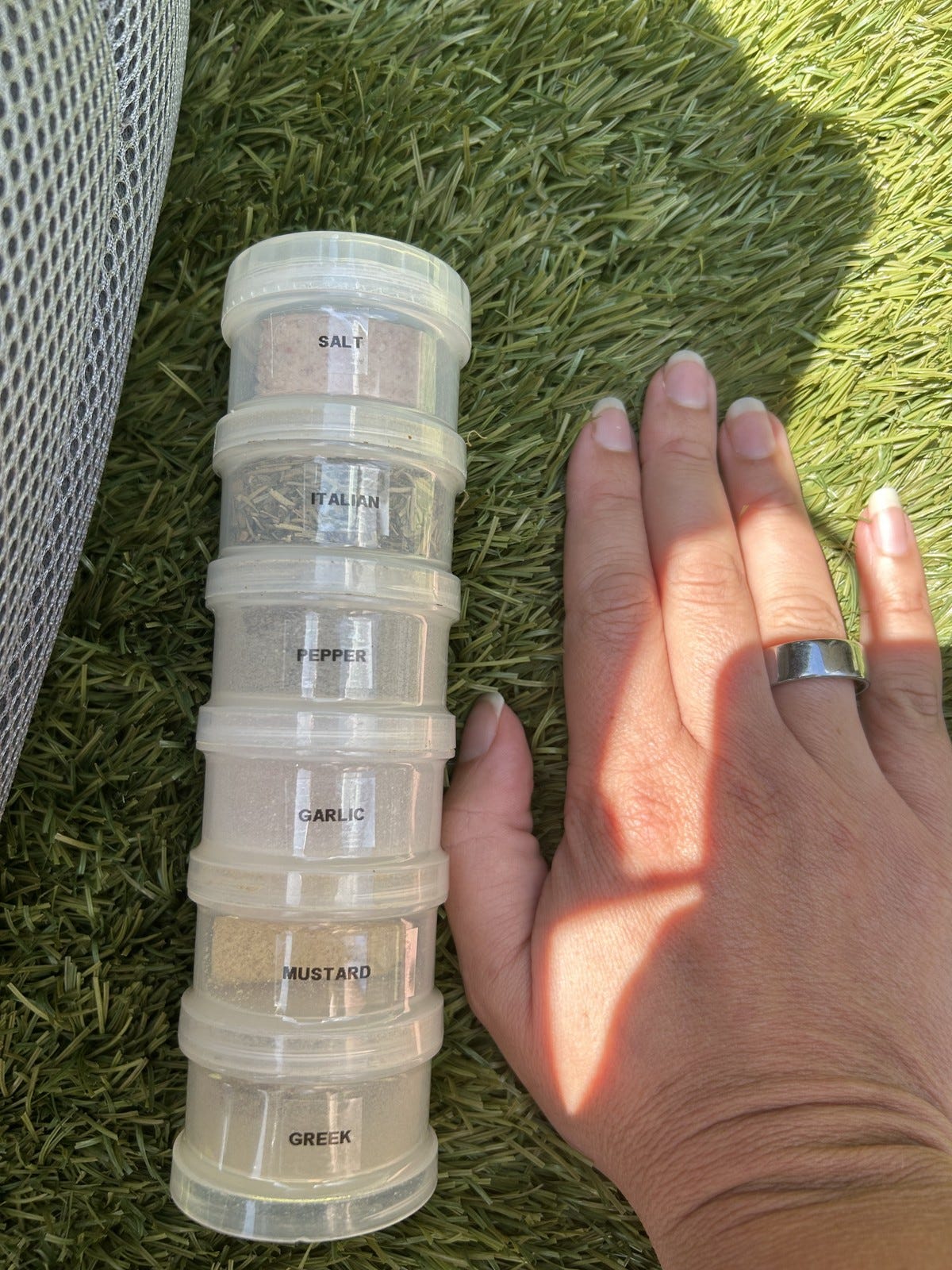 A stackable set of small spice containers labeled Salt, Italian, Pepper, Garlic, Mustard, and Greek, with a hand beside it for scale.