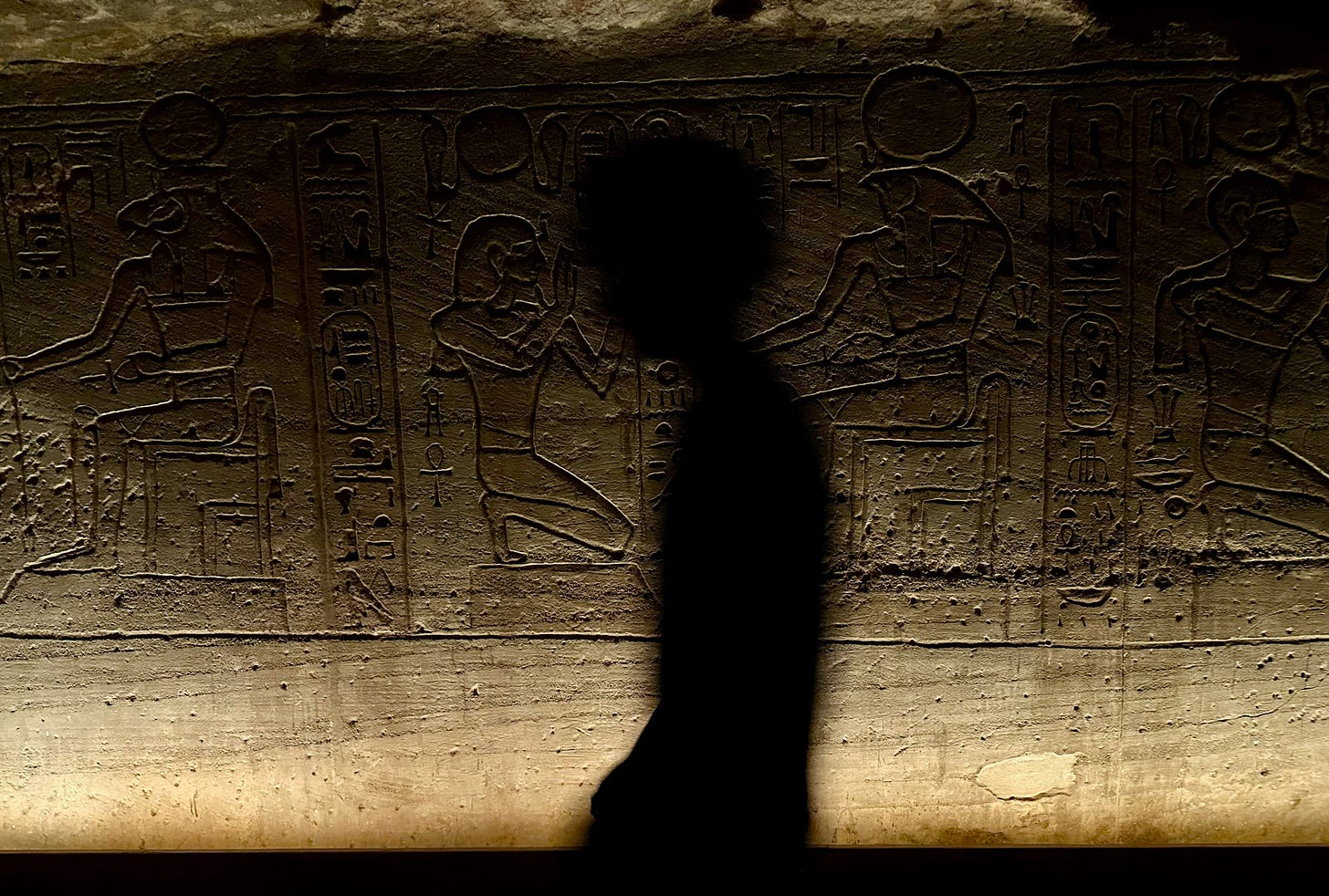 A silhouette of a figure walking past carved hieroglyphics and reliefs inside the Great Temple at Abu Simbel, lit from below
