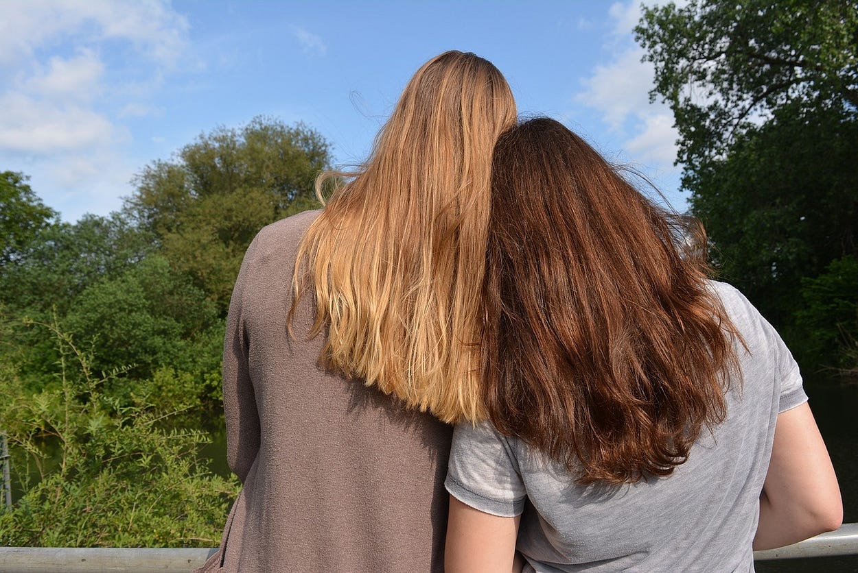 Image shows two woman from the back, one blond and taller thn the brunette, who leans her head on the blonde’s shoulder.