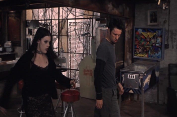 Screen capture from the movie BOOK OF SHADOWS: BLAIR WITCH 2 featuring a goth woman in all black and a skinny man looking down at something off camera. In the background, the pinball machine TIME WARP stands against the wall.