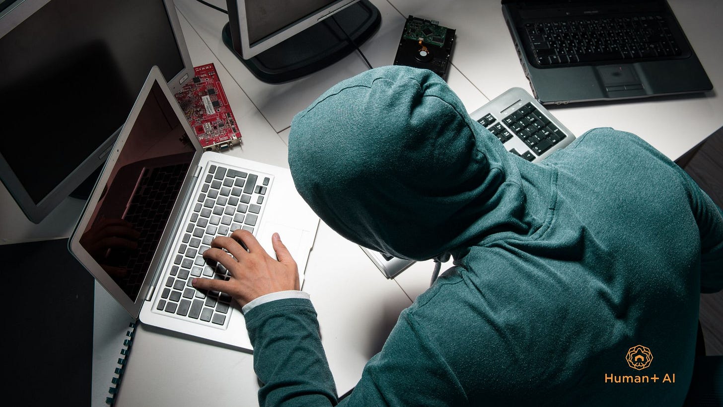 A person wearing a teal hoodie hunches over a laptop surrounded by computer monitors.