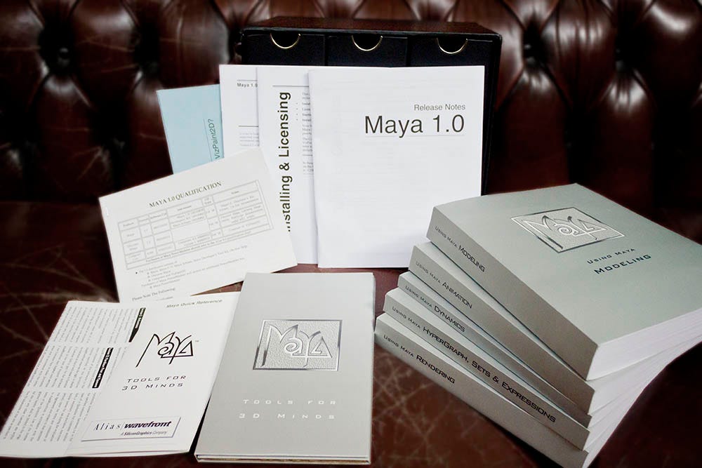 The contents of an original Maya 1.0 software package laid out on a leather couch, including the Release Notes booklet, Installing and Licensing guide, Quick Reference, 'Tools for 3D Minds' booklet, and a stack of silver Maya Library manuals (Modeling, Animation, Dynamics, Hypergraph Sets and Expressions, Rendering)