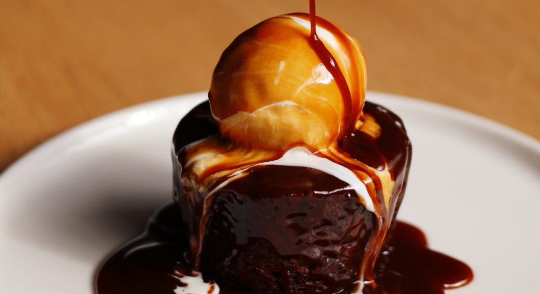A white plate with sticky toffee pudding with ice cream on top and sauce being poured on top of it