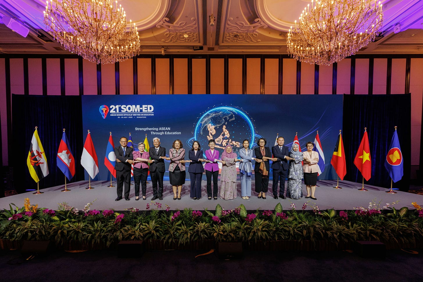 Twelve official delegates, comprising men and women in formal attire, stand in a row on stage performing the traditional ASEAN cross-arm handshake. The stage backdrop features a large blue screen reading "21st SOM-ED: Strengthening ASEAN Through Education", flanked by a row of ASEAN member state flags. Twelve official delegates, comprising men and women in formal attire, stand in a row on stage performing the traditional ASEAN cross-arm handshake. The stage backdrop features a large blue screen reading "21st SOM-ED: Strengthening ASEAN Through Education", flanked by a row of ASEAN member state flags.