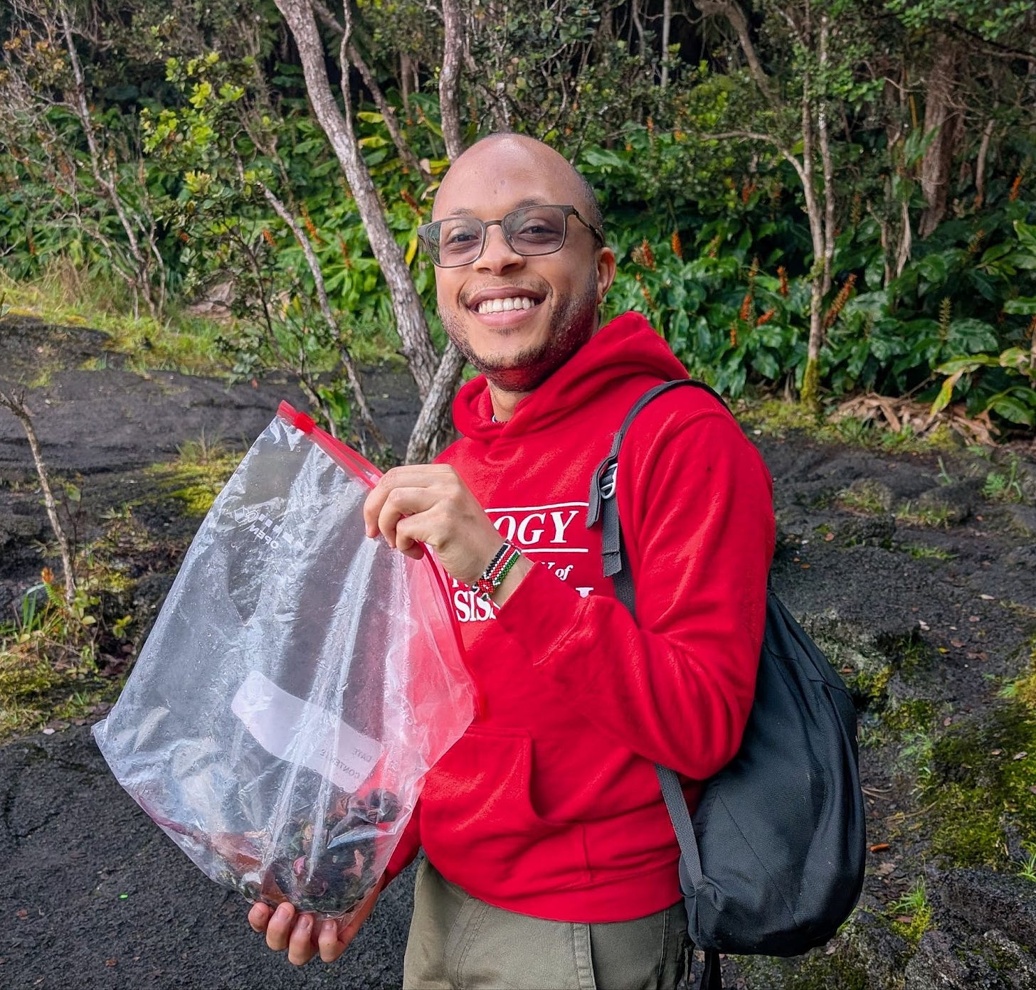 A student with a red UM Biology hoodie, standing in the middle of a nature area with a plastic bag of organic material