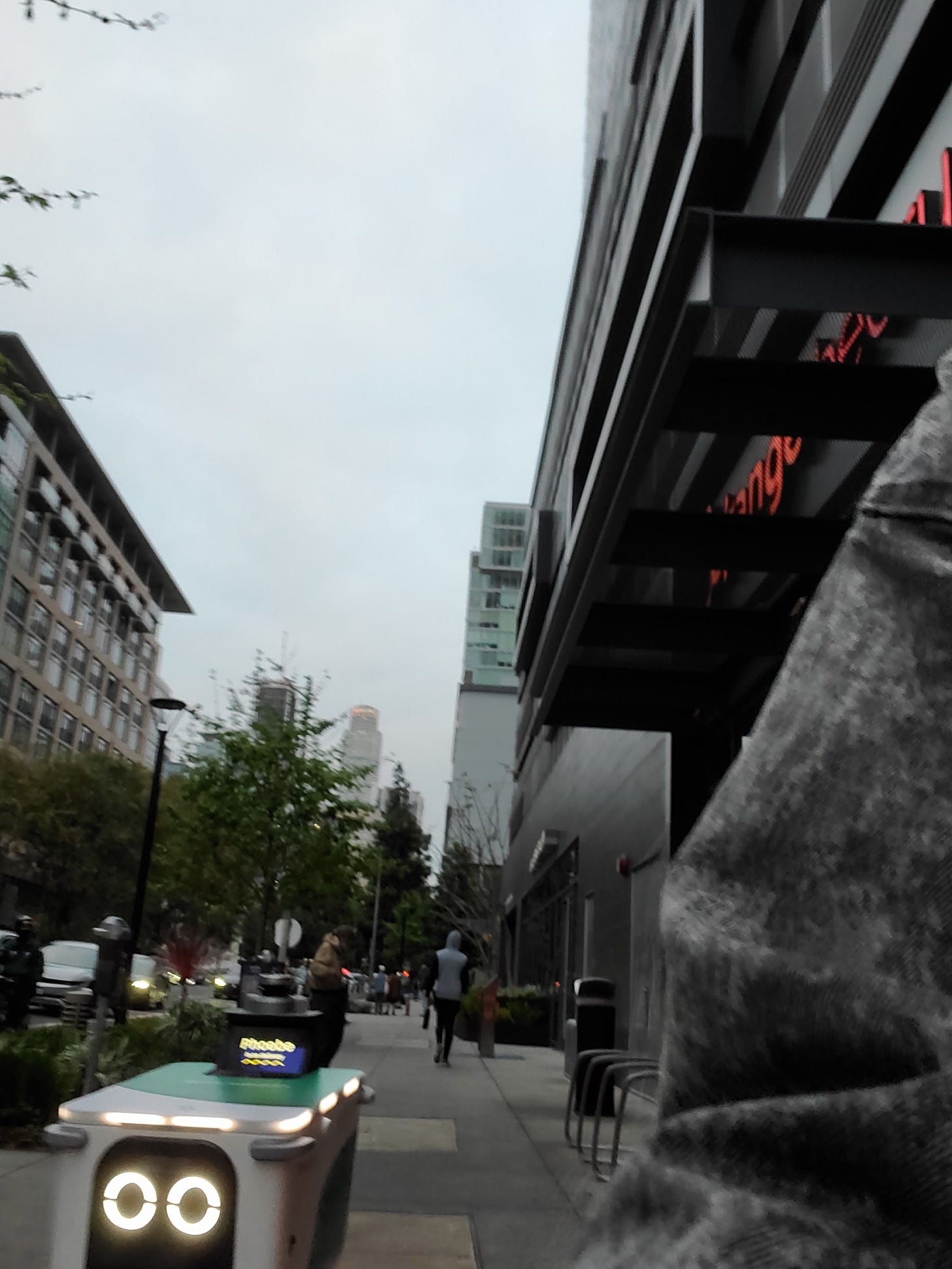 A small robot with haunting LED eyes and a nametag reading "Phoebe" stalks a photographer down a gray city street. A small robot with haunting LED eyes and a nametag reading "Phoebe" stalks a photographer down a gray city street.
