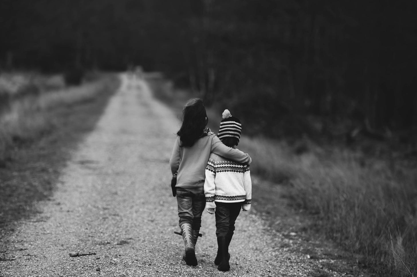 A black and white photo of two children walking on a gravel road, their backs to the camera. One child is putting their arm around the other. A black and white photo of two children walking on a gravel road, their backs to the camera. One child is putting their arm around the other.
