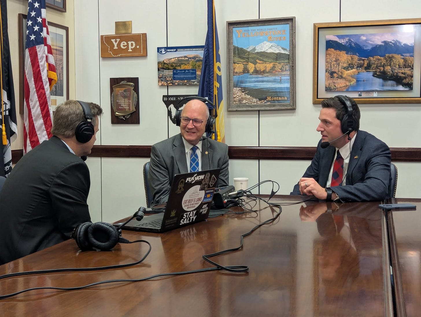 three men in suits recording a podcast at a wooden table inside three men in suits recording a podcast at a wooden table inside