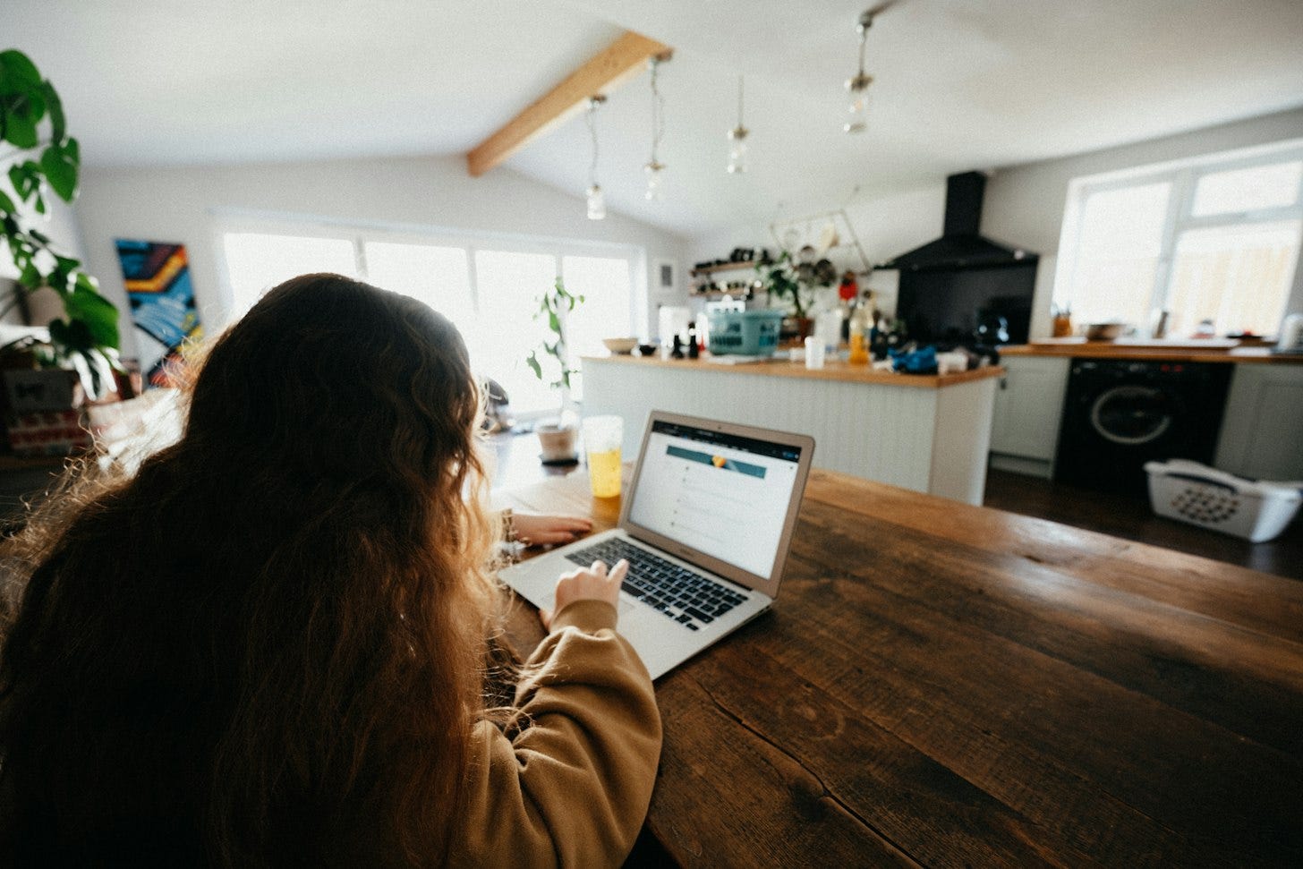 A person with long hair, seen from behind, using a laptop with a web page open at a wooden kitchen table in a bright, sunlit home. A person with long hair, seen from behind, using a laptop with a web page open at a wooden kitchen table in a bright, sunlit home.