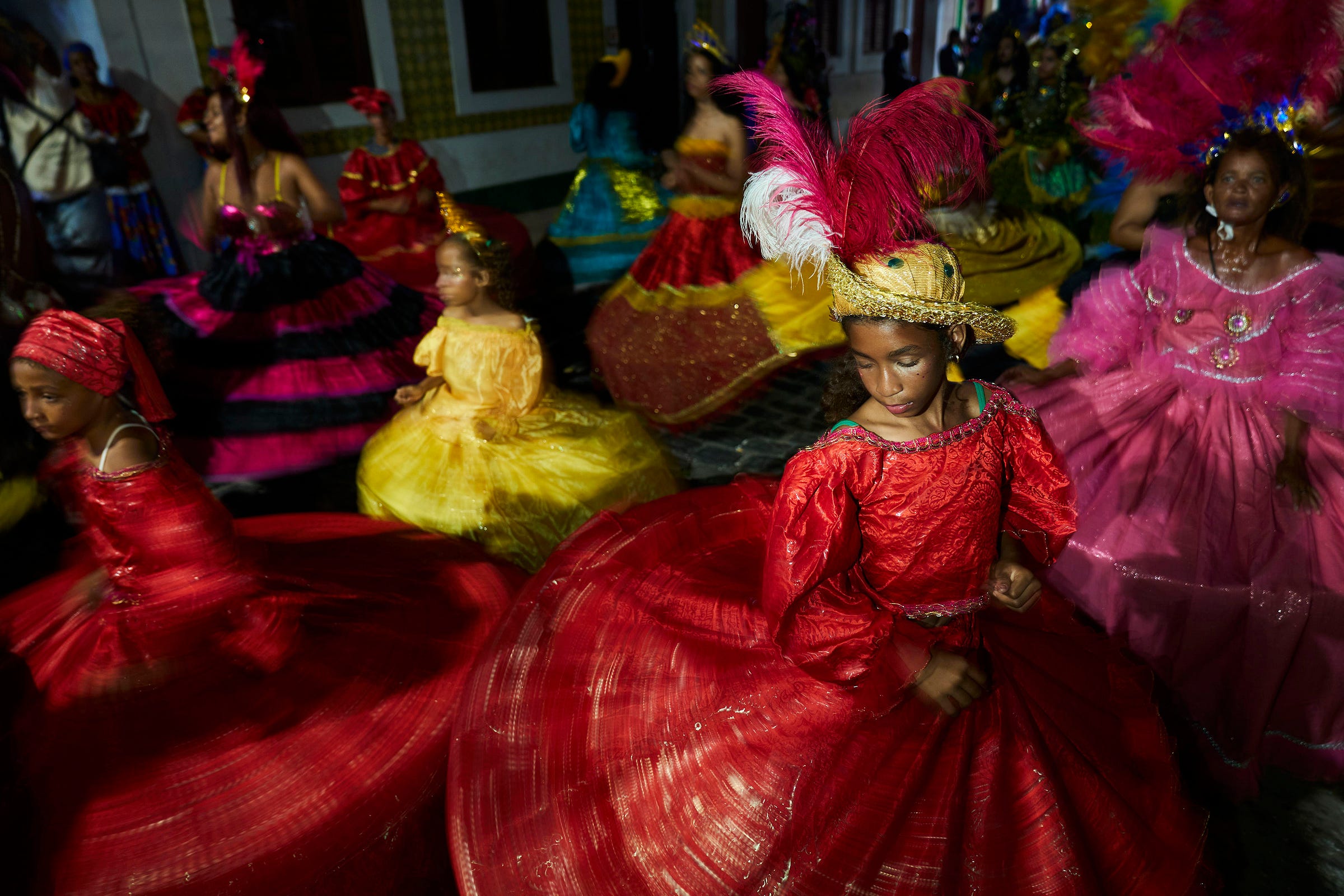 Girls in vibrant, colorful dresses dance in a lively, festive setting. The girl in the foreground wears a red dress with a feathered headdress, while others in yellow, pink, and multicolored dresses move around her.