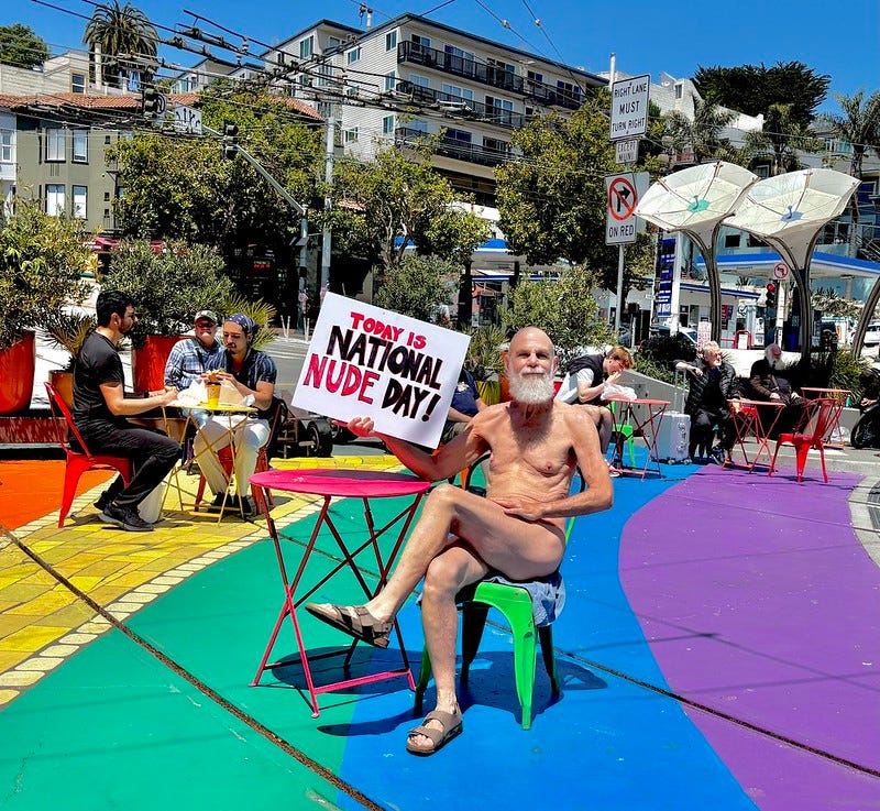 A nude man with a white beard sits on a green chair in Jane Warner Plaza, holding a sign that reads “Today is National Nude Day!” The rainbow-colored pavement and outdoor café tables are visible around him.