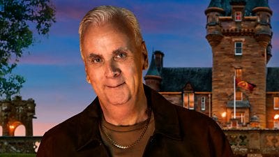 A man in a dark jacket and brown t-shirt smiles to camera, set against the backdrop of a castle by night A man in a dark jacket and brown t-shirt smiles to camera, set against the backdrop of a castle by night