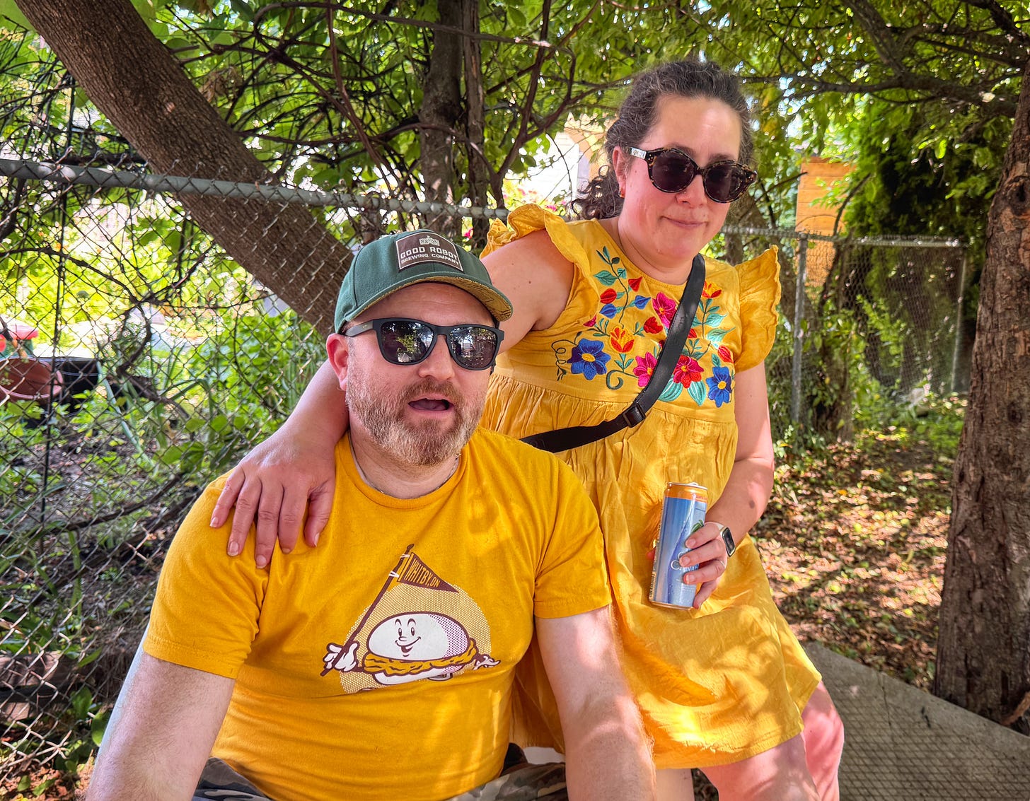 A couple, sitting at the tasting, both wearing mustard-colour clothing. The man is wearing a mustard-coloured t-shirt with an illustration of a hamburger on it. The woman is wearing a mustard-coloured sun dress with contrasting access colours.