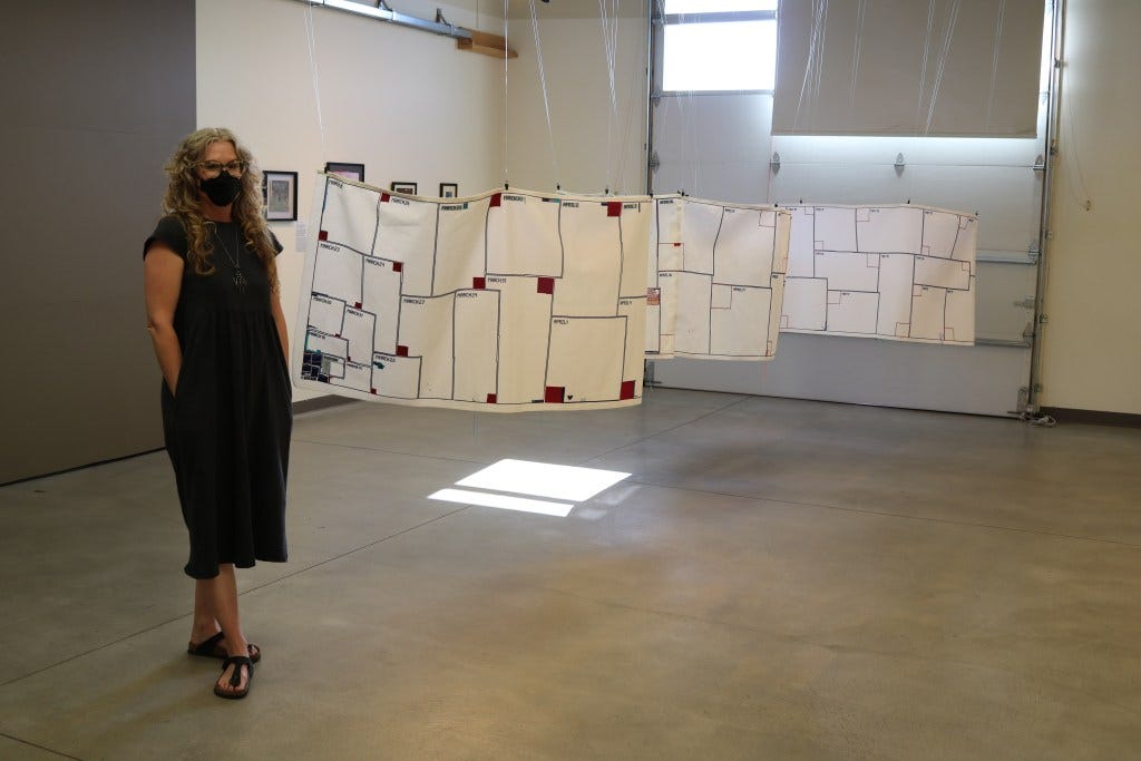 A white femme poses beside a panel from a tapestry with colored boxes representing COVID-19 cases and deaths. She wears a black mask that matches her black dress.