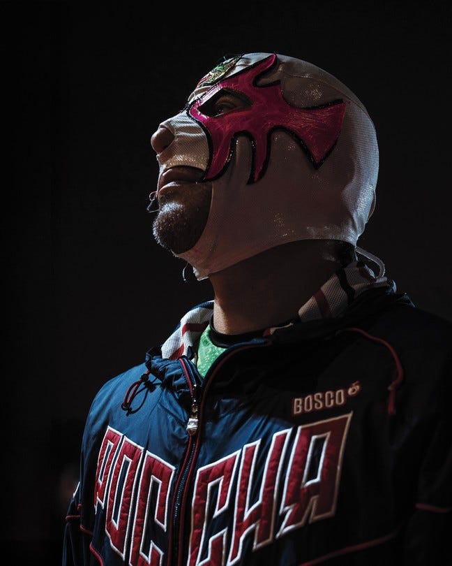 A color photograph of a man wearing a skin-tight white-and-pink luchador mask, set against a black background.