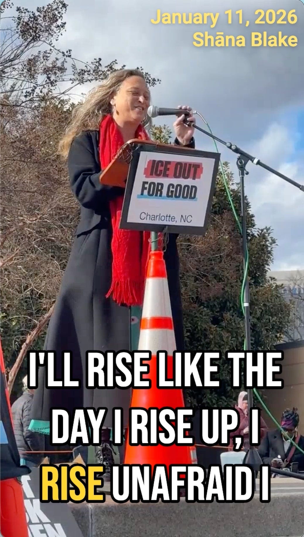  A woman, identified as Shāna Blake, speaks at a podium outdoors in Charlotte, NC, on January 11, 2026. A sign on the podium reads "ICE OUT FOR GOOD," and large overlay text at the bottom features lyrics: "I'LL RISE LIKE THE DAY I RISE UP, I RISE UNAFRAID I."