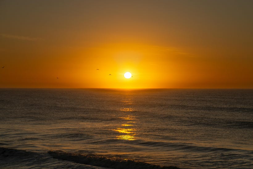 A sunrise over an ocean. The image is divided in half by the horizon line. The sun is already a little above the water, but the sky is still orangey-yellow, with the light rippling over calm seas. A sunrise over an ocean. The image is divided in half by the horizon line. The sun is already a little above the water, but the sky is still orangey-yellow, with the light rippling over calm seas.