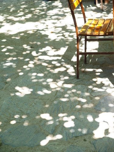 a photo of a wooden chair and the ground beneath the chair full of the shadows of tree leaves aka dappled sunlight