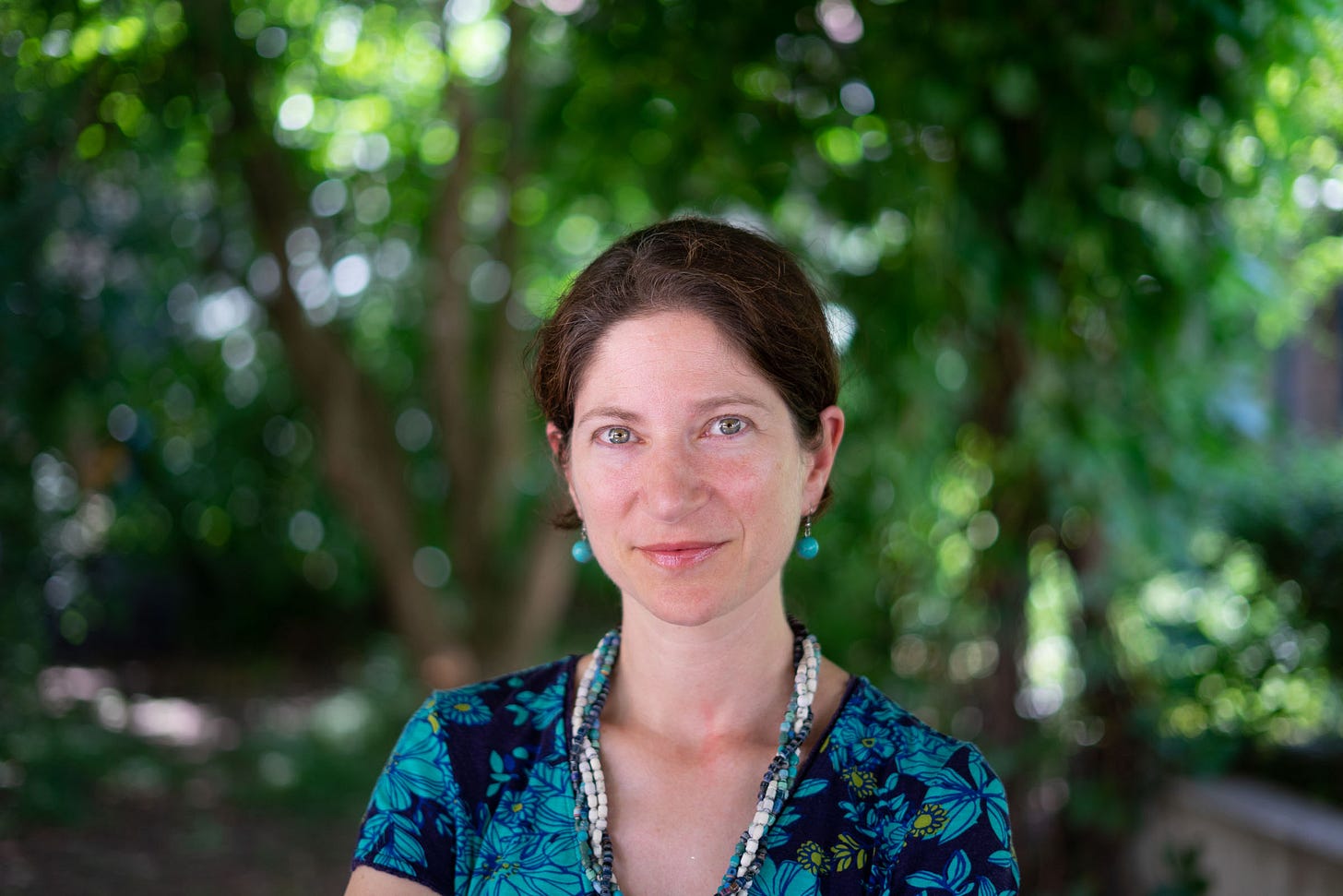 Picture of Julia Steinberger in a green outdoor space, looking directly at the camera. She has brown hair and is wearing blue earrings, a blue & white necklace and blue & black dress