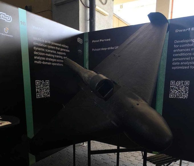 A black pulsejet kamikaze drone, KROOK-1, displayed on a stand. The drone has a sleek, triangular design with a pointed nose and visible engine intake. Surrounding panels feature text and QR codes, including references to First Parsec and IT Arena. A black pulsejet kamikaze drone, KROOK-1, displayed on a stand. The drone has a sleek, triangular design with a pointed nose and visible engine intake. Surrounding panels feature text and QR codes, including references to First Parsec and IT Arena.