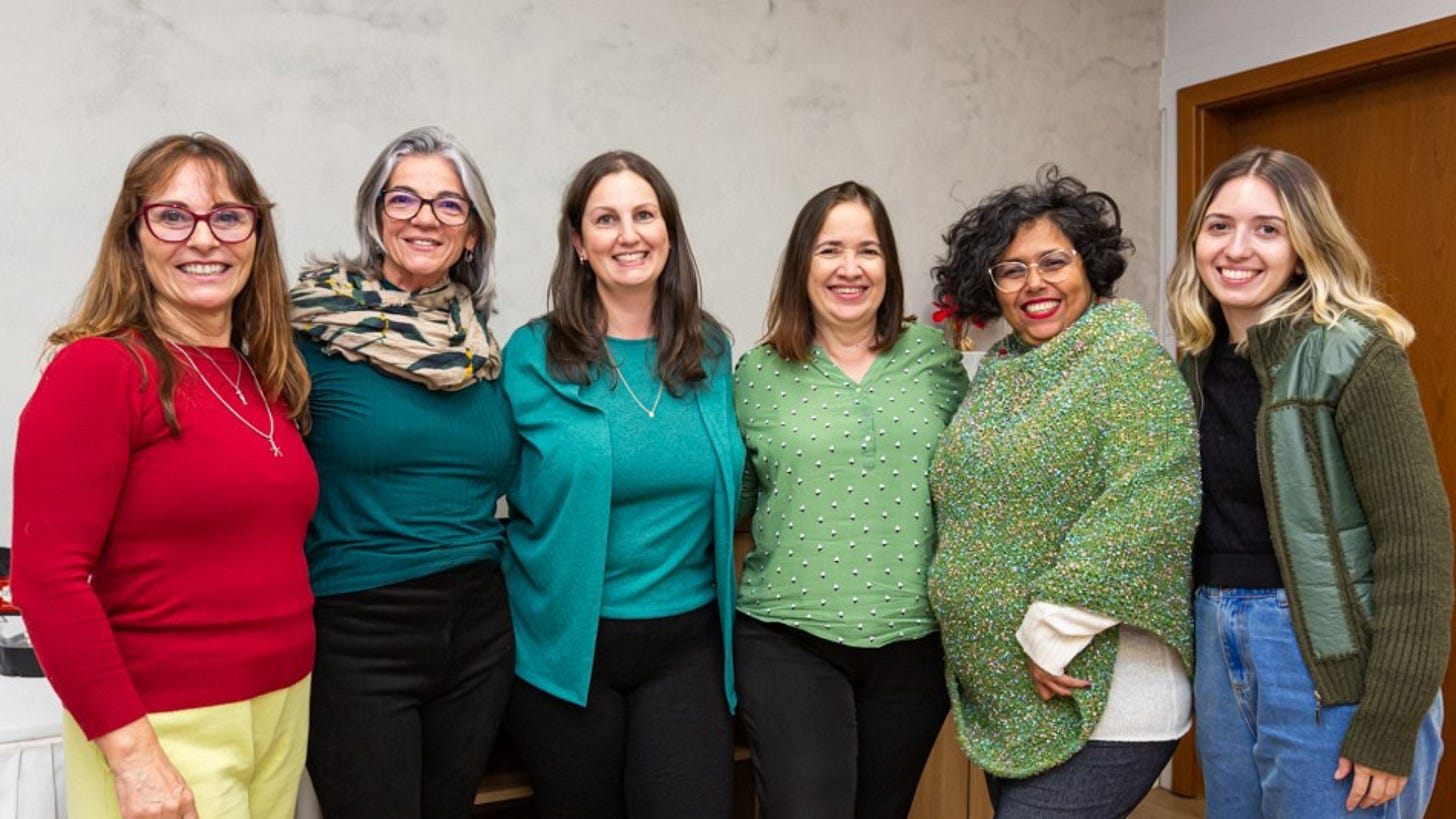 #PraTodosVerem: Da esquerda para a direita, Cristina Rabelais, mulher branca de cabelos castanho-claro e compridos, usa óculos de grau, blusa de manga comprida vermelha e calça amarela. Ao lado, Maria Fernanda Bittencourt, mulher branca de cabelos grisalhos e compridos, usa óculos de grau, um xale estampado em bege, verde, azul e amarelo, blusa verde e calça preta. Em seguida, Mariana Gabriel, mulher branca de cabelos castanhos e compridos, veste uma blusa verde, cardigan verde e calça preta. Ao seu lado, Dolores Abreu, mulher branca de cabelos castanhos na altura dos ombros, usa uma blusa verde com bolinhas brancas e calça preta. Depois, Silvia Pereira, mulher negra de cabelos curtos, pretos e cacheados, usa um poncho verde com bolinhas coloridas, uma blusa branca por baixo e calça cinza escura. Por fim, Daniele Novaes, mulher branca de cabelos loiros descoloridos nas pontas, usa uma blusa preta, casaco verde e calça jeans. Ao fundo, uma parede cinza e uma porta. Fim da descrição.