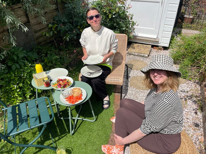L-R: two white women sit in a garden, smiling at the camera, it's very sunny, there's food on the table between them, a mandala on the forest floor with a candle, flowers and cards, trees in the forest, three crocuses, two white, one purple