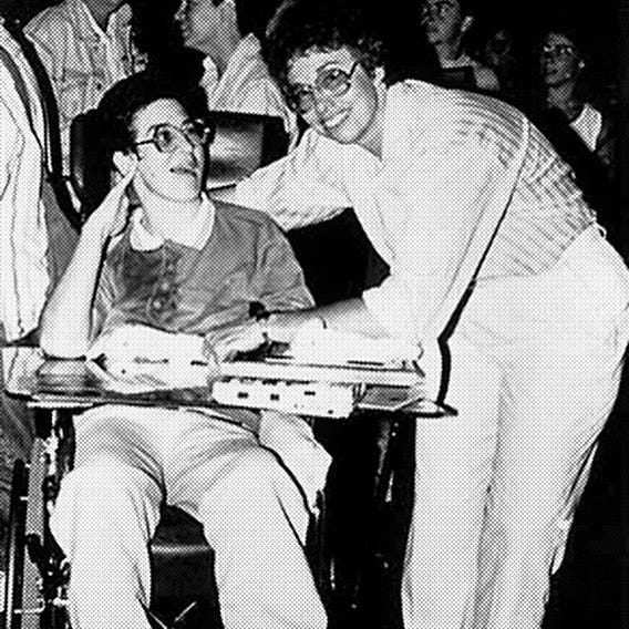 In a grainy black and white photo, two white lesbians. One is hunched over the desk attached to her partner’s powerchair. In a grainy black and white photo, two white lesbians. One is hunched over the desk attached to her partner’s powerchair.