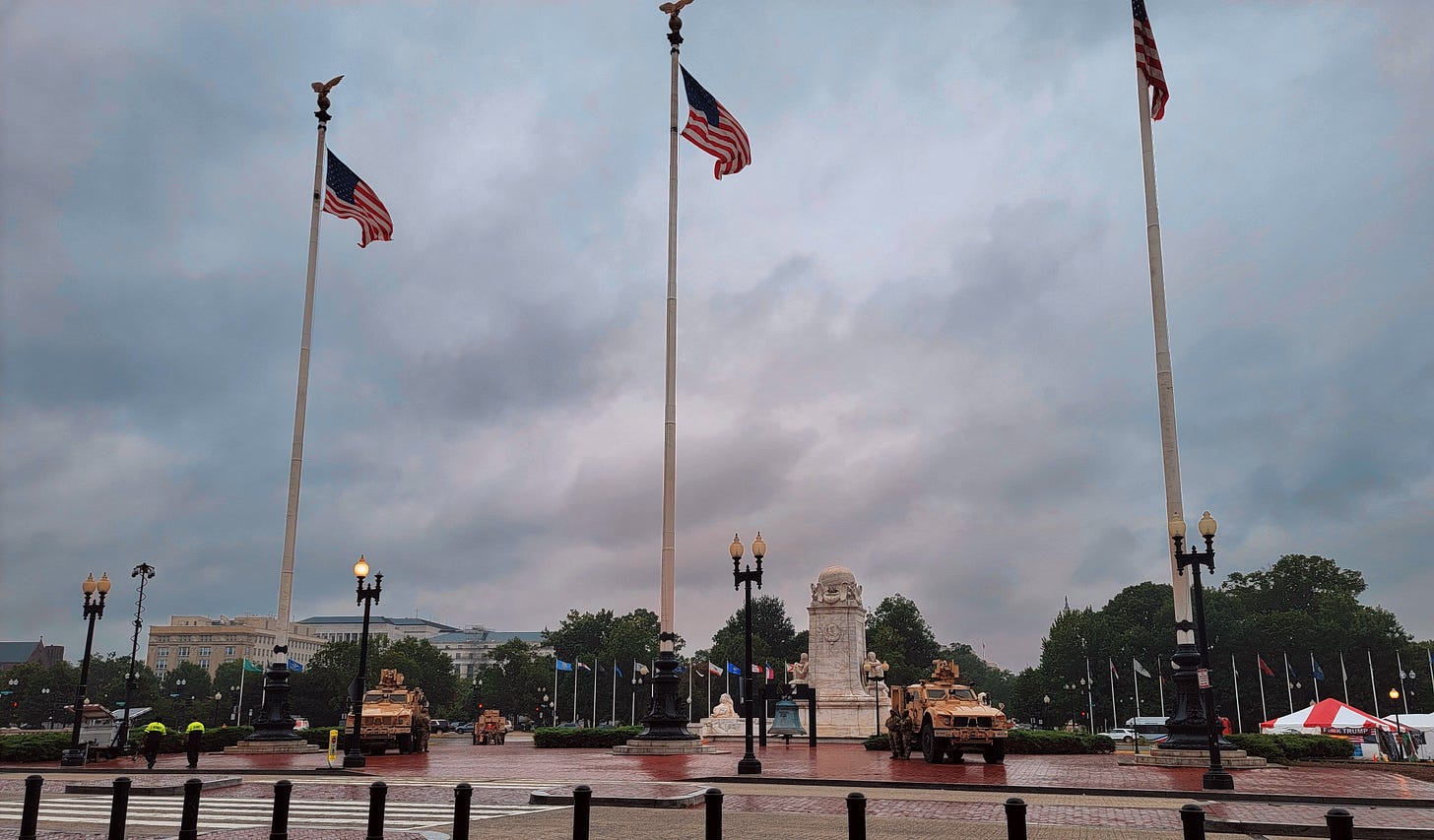 Two large military vehicles parked in the gaps between three flagpoles, each flying the American flag. They're in a brick plaza across the street from something (it's Union Station, but you can't see that from the pic). A green-tarnished copper bell hangs behind them from some kind of steel girder, and there's a square marble piece of statuary behind that. In the distance, some official-looking marble building and green trees