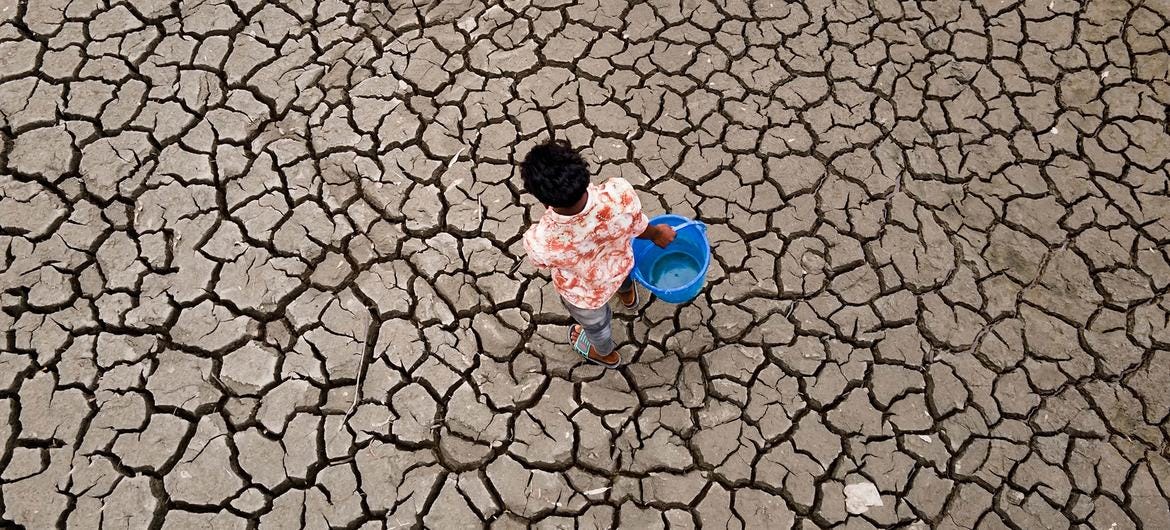 Overhead view of a young man carrying a blue bucket across a vast expanse of cracked, dry earth in Bangladesh. Overhead view of a young man carrying a blue bucket across a vast expanse of cracked, dry earth in Bangladesh.