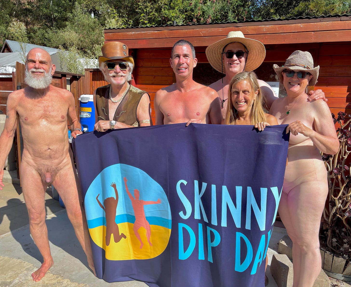 Six nude adults holding a Skinny Dip Day banner pose in front of a small wooden building. The group is smiling and enjoying the sun during a celebratory naturist event in California. Six nude adults holding a Skinny Dip Day banner pose in front of a small wooden building. The group is smiling and enjoying the sun during a celebratory naturist event in California.