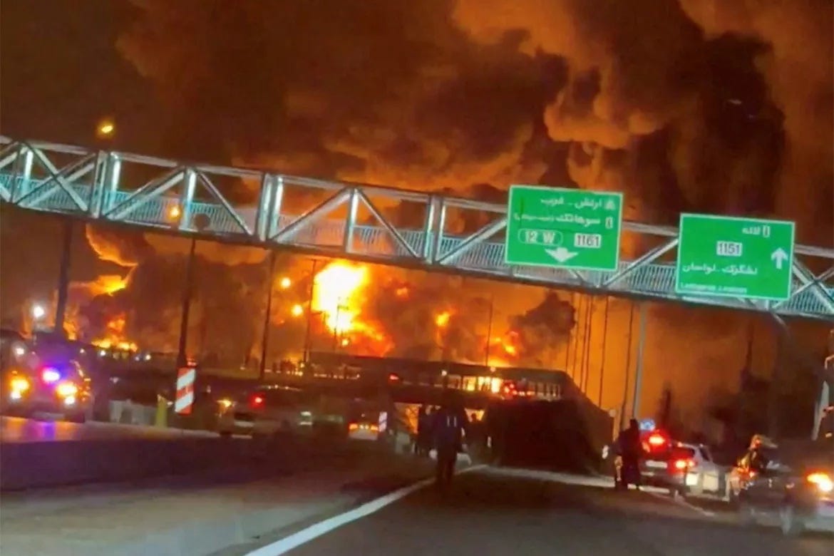 A massive fire burns at an oil facility on the outskirts of Tehran, Iran. Emergency vehicles and stopped traffic line a motorway as thick black smoke and orange flames fill the night sky. Persian-language road signs are visible overhead. The Straits of Hormuz. The price going up. This is what it looks like when the chain tightens.